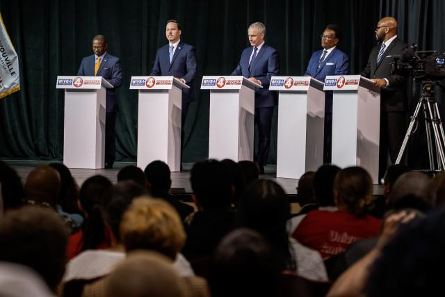 View of 5 men behind podiums standing on a stage from the audience of a theatre.