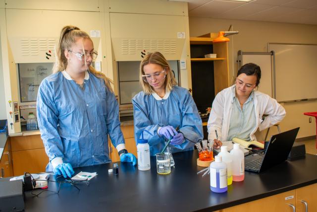 Three women stand in a lab at a table. One woman is using a pipette to move liquid into a beaker.