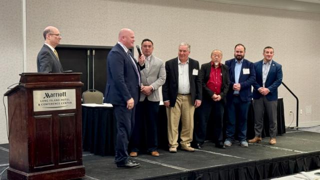 A group of people stand in a line on a stage. One person is talking into a microphone. 
