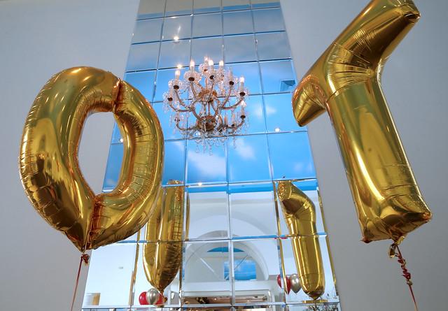 Two gold foil balloons in the shape of the letters "O" and "T" float in front of a large, paneled mirror. A chandelier hangs above them.