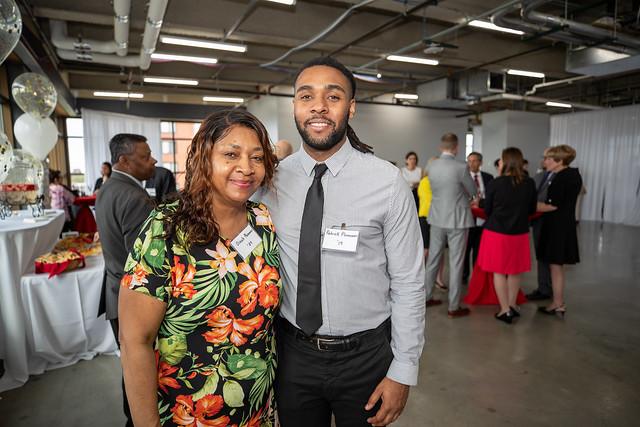 A young, African American man stands with an older African American woman while a party goes on in the background. Both of them are smiling and wearing professional-looking clothes and nametags.
