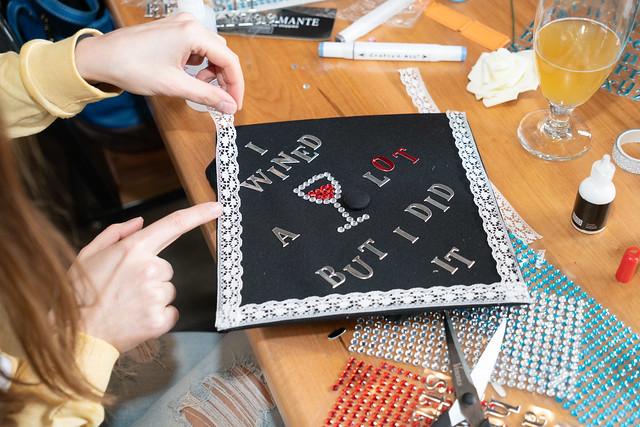 Close-up of a girl's hands applying lace to a decorated mortorboard. The text on the mortarboard says "I WINED a Lot but I did it!", and is accompanied by a picture of a wine glass.