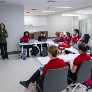 A group of nursing students attend lab class. Students sit in U shape facing a screen while professor lectures.