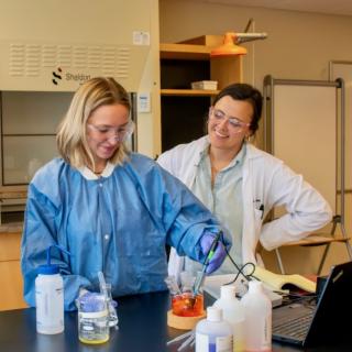 Three women stand in a lab at a table. One woman is using a pipette to move liquid into a beaker.