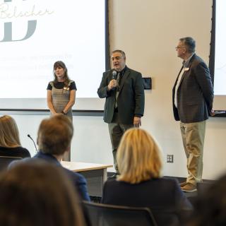 three people stand on stage addressing an audience. The man in the center stands with a microphone.