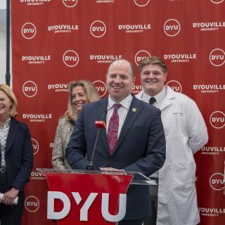 man smiles and stands at a podium that reads DYU while addressing the crowd