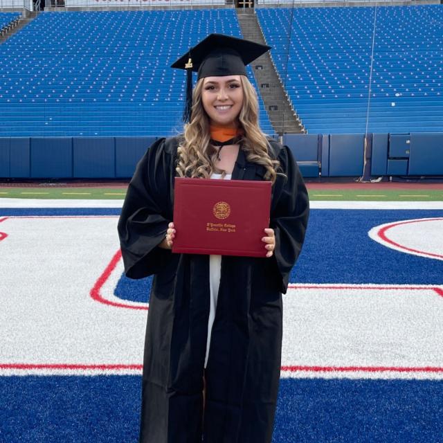 Rachel Rainey, Woman smiles at the camera wearing graduation regalia holding up a D'Youville University Diploma.