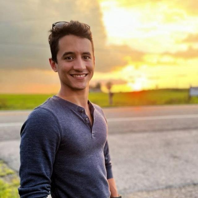 Giovanni Feliciano, man with dark hair wearing a blue shirt smiles at the camera in front of a sunset.