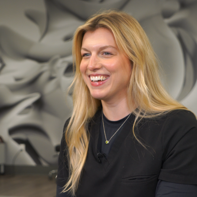 Blond woman smiles at the camera. She wears scrubs and sits in a large room in front of a spine wall mural